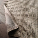 A close-up of a textured area rug in a warm taupe-beige color, shown with one corner folded over to reveal the underside, placed on a dark wood floor.