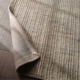 A close-up of a textured area rug in a warm taupe-beige color, shown with one corner folded over to reveal the underside, placed on a dark wood floor.