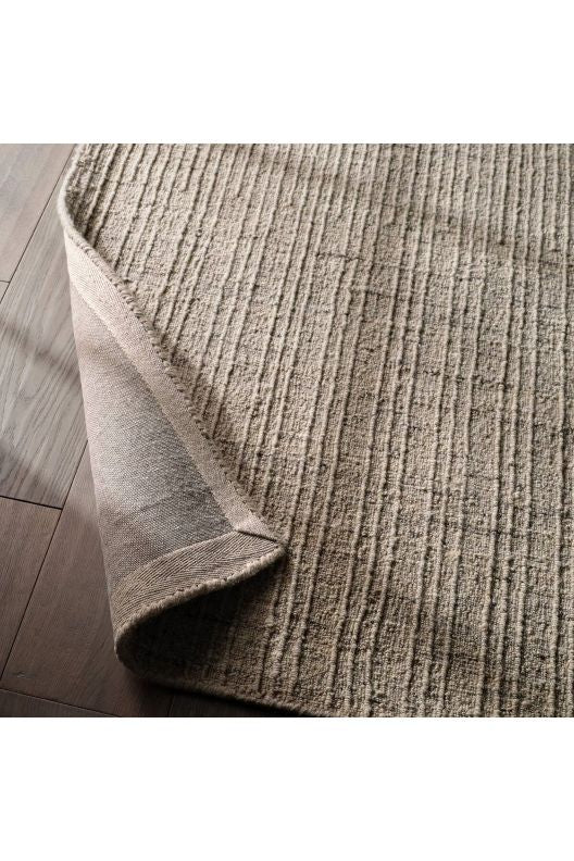 A close-up of a textured area rug in a warm taupe-beige color, shown with one corner folded over to reveal the underside, placed on a dark wood floor.