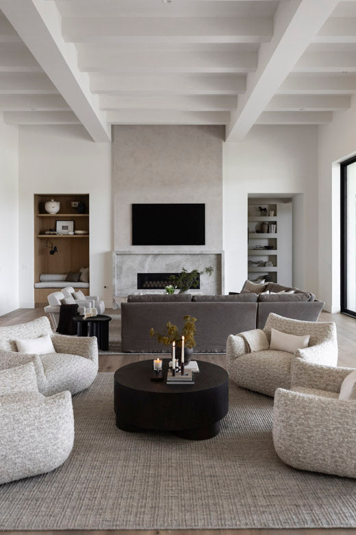 A modern living room with a large round black coffee table surrounded by four textured light beige armchairs on a woven gray and cream area rug. In the background, a gray sectional sofa sits in front of a fireplace with a mounted TV above it, under a white coffered ceiling.