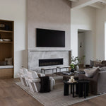 A modern living room interior featuring a large black flat-screen TV mounted above a stone fireplace. The room has light wood floors, a textured gray and cream area rug, a gray sofa with pillows, white accent chairs, and a built-in wooden shelf on the left.