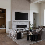 A modern living room interior featuring a large black flat-screen TV mounted above a stone fireplace. The room has light wood floors, a textured gray and cream area rug, a gray sofa with pillows, white accent chairs, and a built-in wooden shelf on the left.
