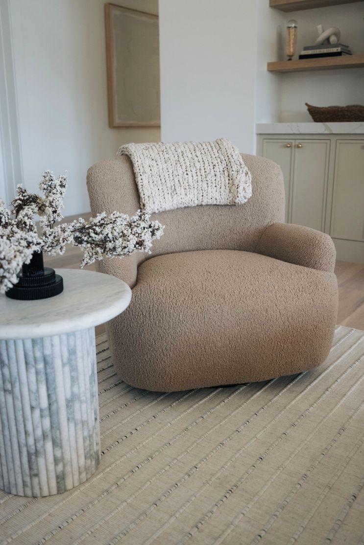 Beige Kadon Swivel Chair upholstered in shearling with softly rolled arms and 360-degree rotation in a living room setting with a side table and decorative elements.