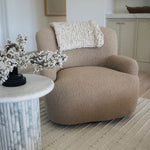Beige Kadon Swivel Chair upholstered in shearling with softly rolled arms and 360-degree rotation in a living room setting with a side table and decorative elements.