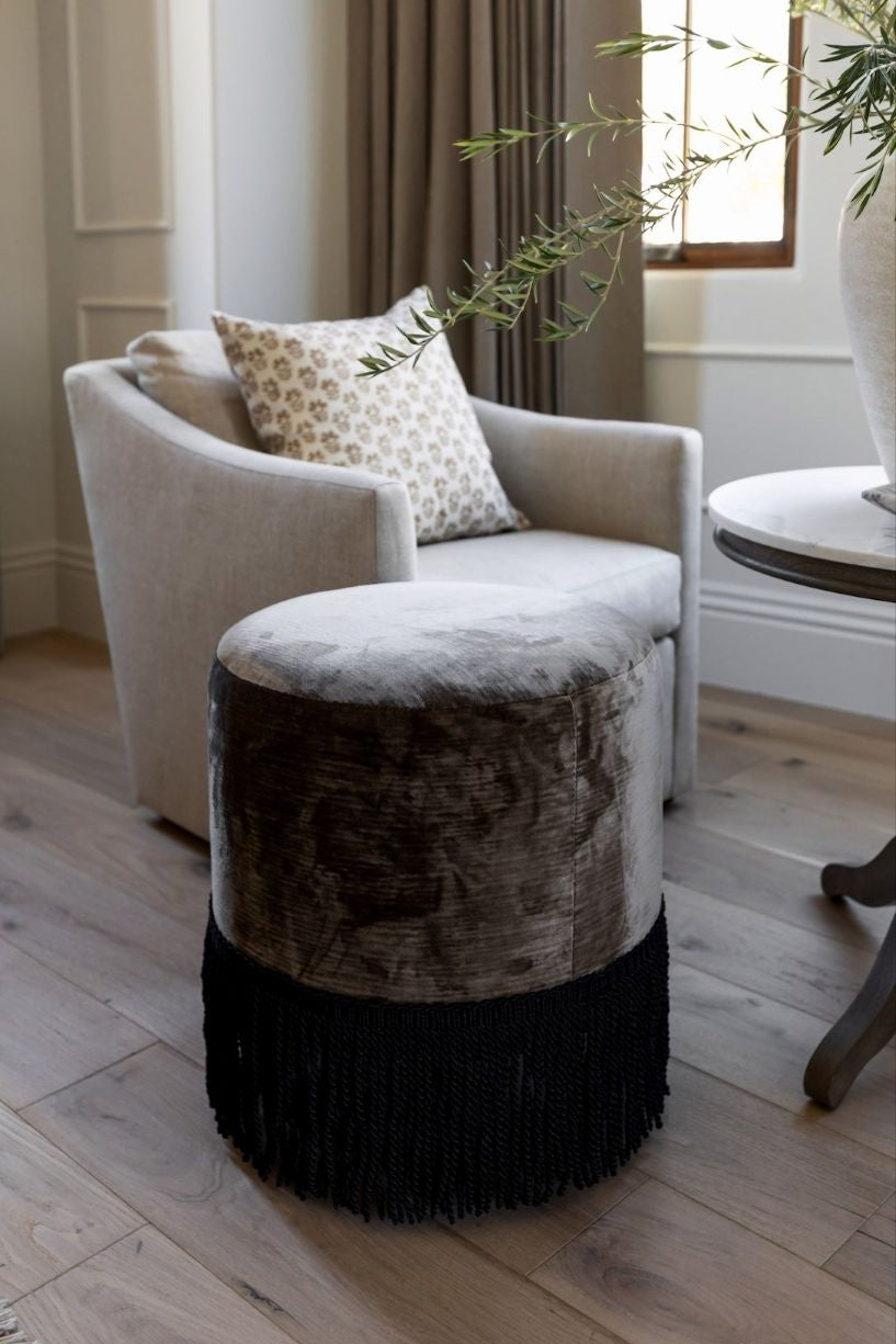 An upholstered ottoman with black fringe on the lower part, placed on a wooden floor next to a beige armchair with a decorative pillow.