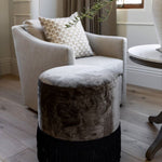 An upholstered ottoman with black fringe on the lower part, placed on a wooden floor next to a beige armchair with a decorative pillow.