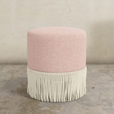 Pink upholstered ottoman with white fringe on a concrete floor and gray wall background