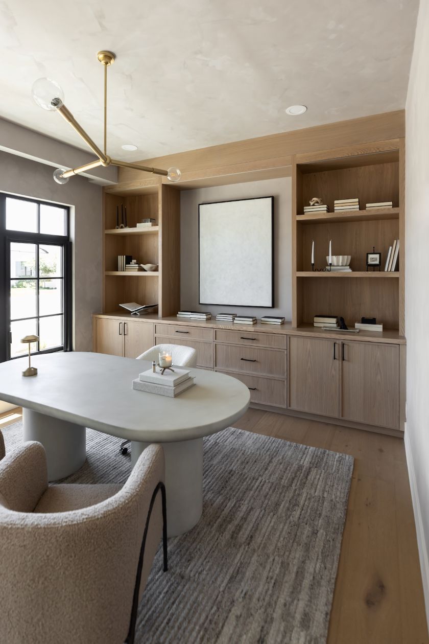 Modern home office with Regina Andrew Talon chandelier, wooden bookshelves, a white desk, and a large window.